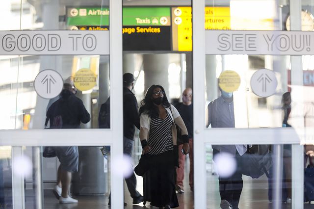 A woman wearing a mask walks through a door at JFK Airport, which says "Good to see you"